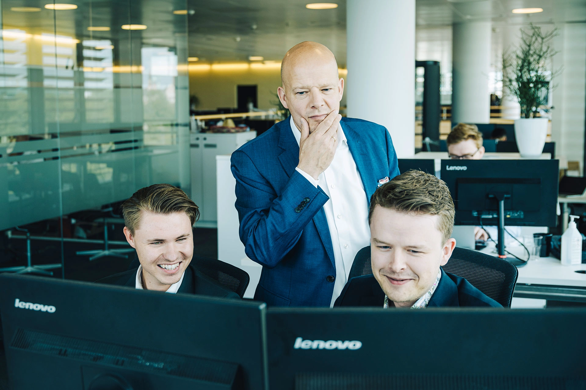 På billedet ses Henrik Wellejus, der betænktsomt tager sig til hagen. Han står i midten af to yngre medarbejdere. Alle tre kigger de på en computerskærm. 