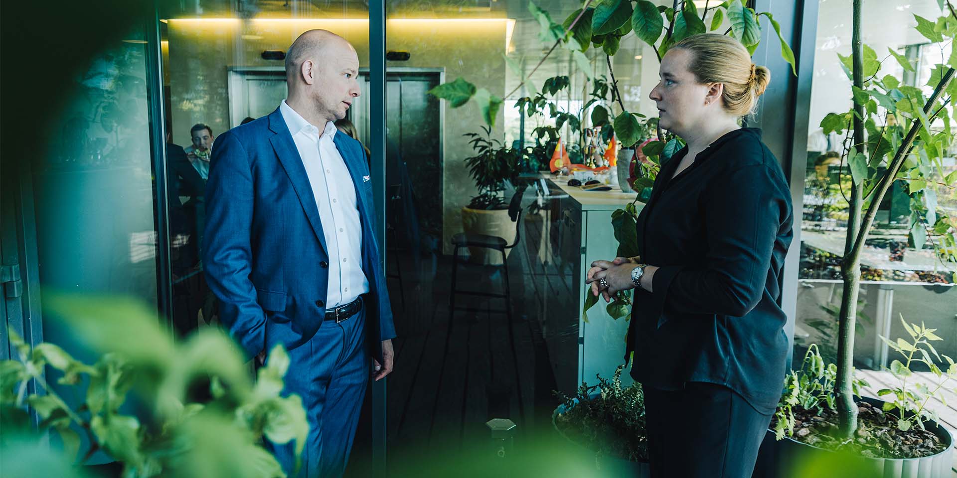 To mennesker står og taler sammen i en kontorlandskab med grønne planter - vi ser dem fra siden. Til venstre står Henrik Wellejus og til højre står assistant manager Emilie Hjort Jensen