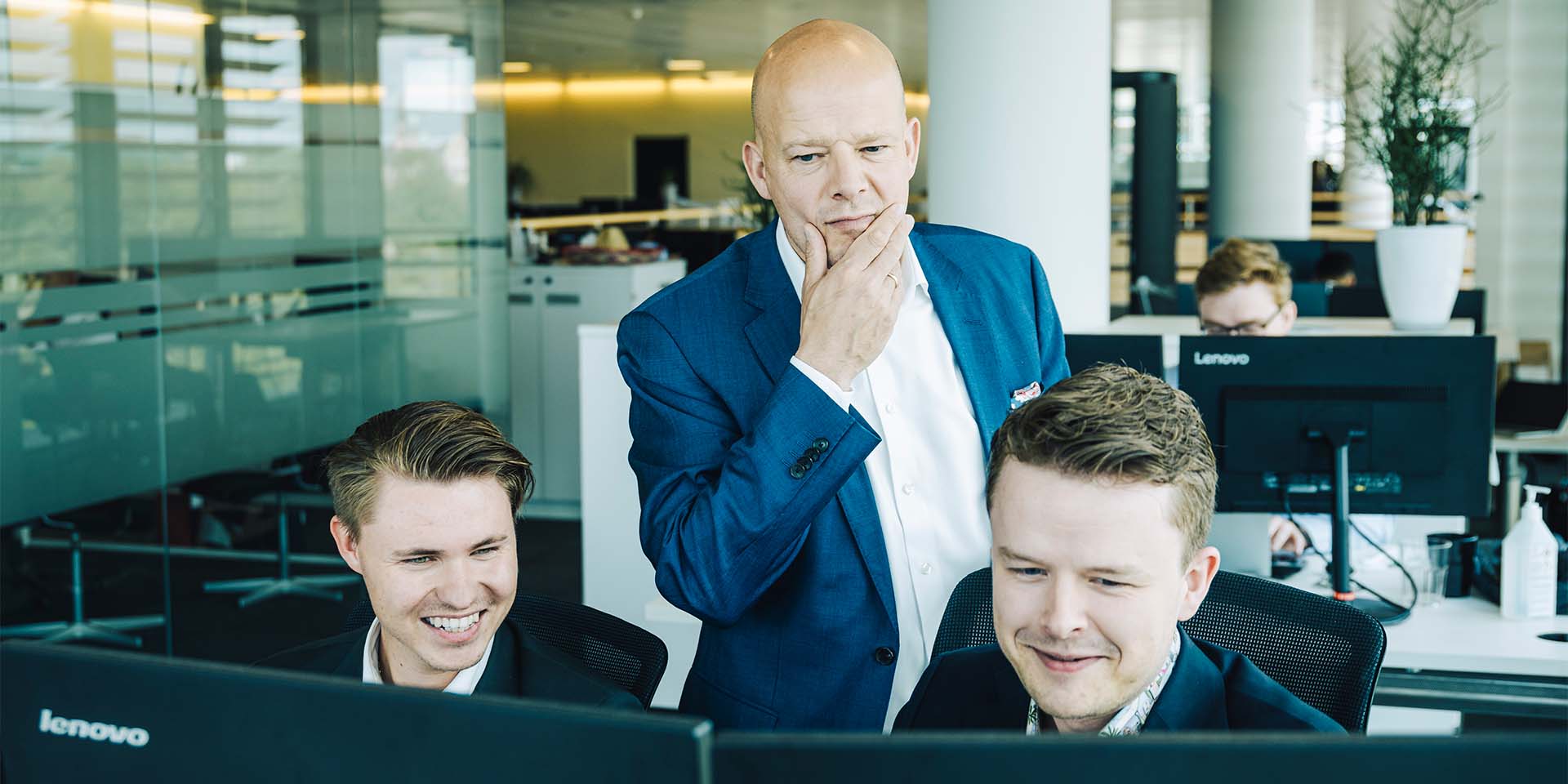 På billedet ses Henrik Wellejus, der betænktsomt tager sig til hagen. Han står i midten af to yngre medarbejdere. Alle tre kigger de på en computerskærm. 